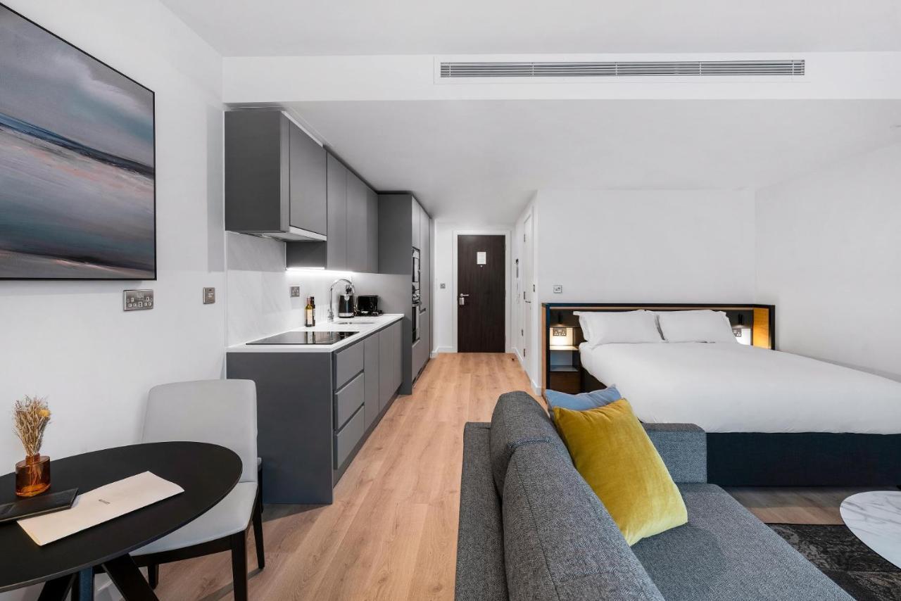 Citysuites 2 Aparthotel Селфорд Екстер'єр фото The photo shows a modern and stylish studio apartment. It features a kitchenette with dark cabinetry and a small dining table with two chairs. There's a cozy sofa with colorful cushions, positioned next to the table. In the background, a large bed wi
