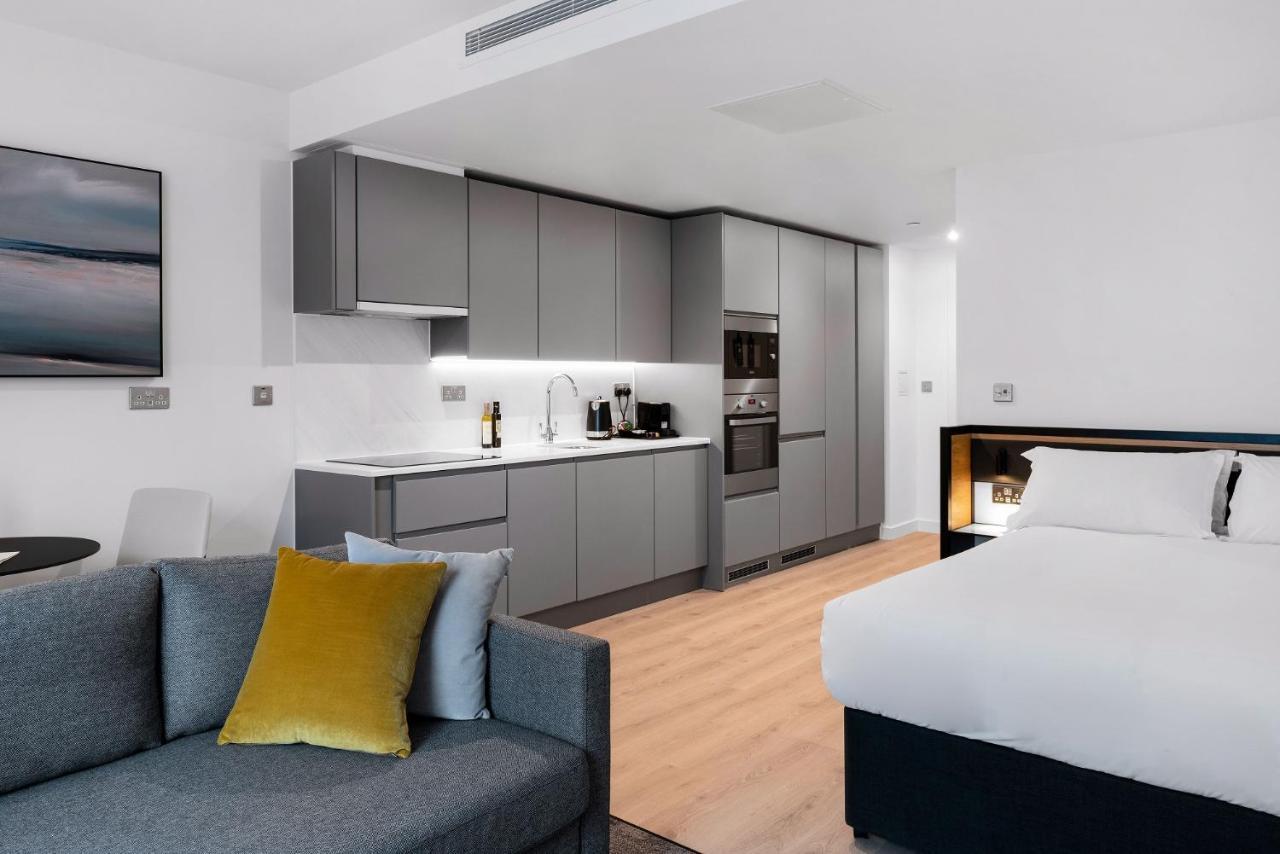 Citysuites 2 Aparthotel Селфорд Екстер'єр фото The photo shows a modern studio apartment featuring an open-plan layout. On one side, there is a kitchenette with sleek, gray cabinets and appliances, including a coffee maker and oven. Adjacent to the kitchenette is a small dining area with a table 