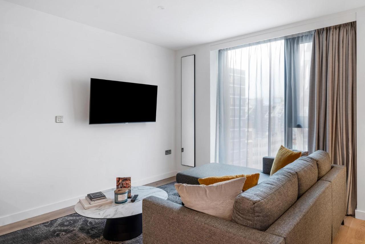 Citysuites 2 Aparthotel Манчестер Екстер'єр фото The image shows a modern living room with a minimalist design. There is a large flat-screen TV mounted on the white wall. A comfortable gray sofa with textured cushions occupies the center of the room, complemented by a round coffee table in front of