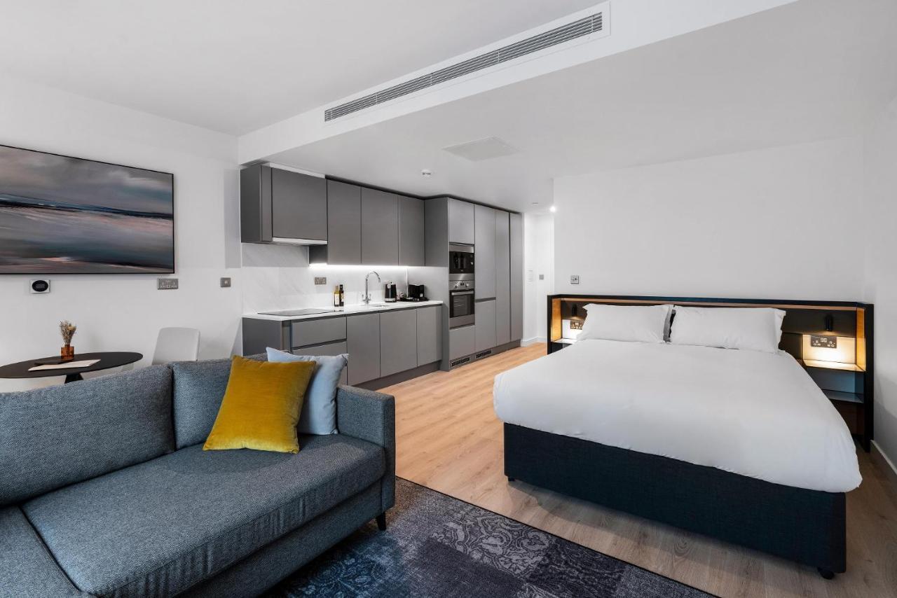 Citysuites 2 Aparthotel Манчестер Екстер'єр фото The photo shows a modern studio apartment. On one side, there is a comfortable gray sofa adorned with a yellow cushion. Nearby, a round table is placed, likely intended for dining or casual sitting. 

The kitchen area features sleek gray cabinetry wi