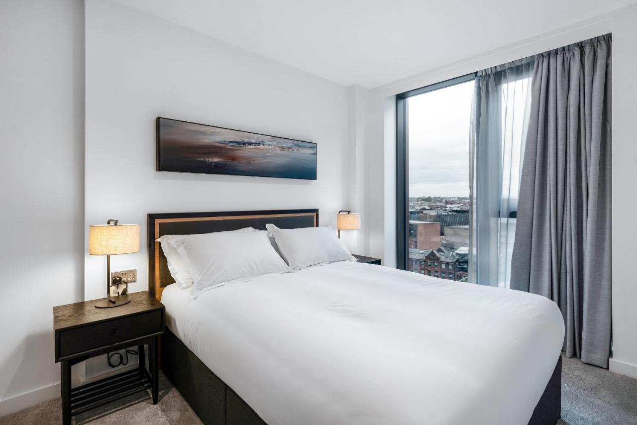 Citysuites 2 Aparthotel Манчестер Екстер'єр фото The image shows a modern bedroom featuring a large bed with a white duvet and two pillows. On either side of the bed are small nightstands with lamps. A large window occupies one wall, draped with light gray curtains, allowing natural light to enter 