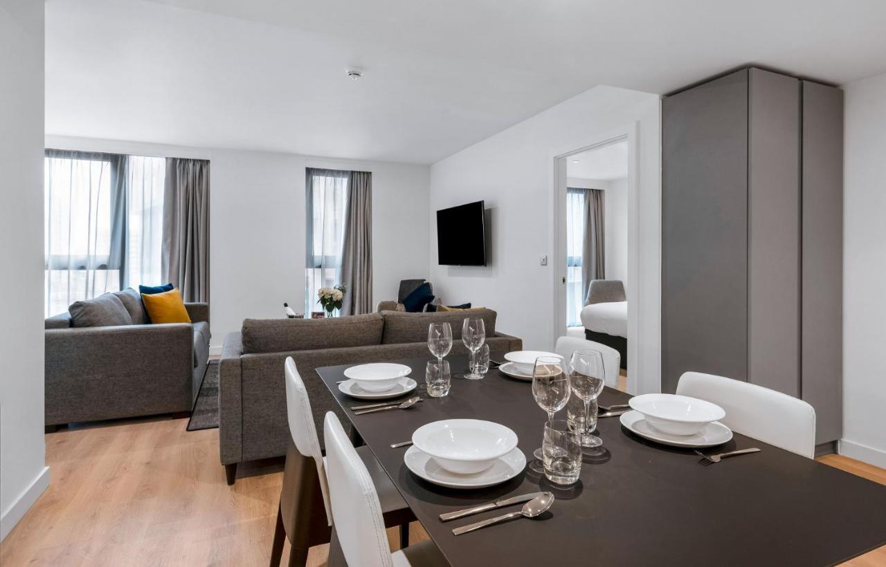 Citysuites 2 Aparthotel Манчестер Екстер'єр фото The photo shows a modern living space with an open layout. In the foreground, there is a dining table set for a meal, featuring multiple plates, glasses, and cutlery arranged neatly. In the background, there is a comfortable-looking gray sofa with a 