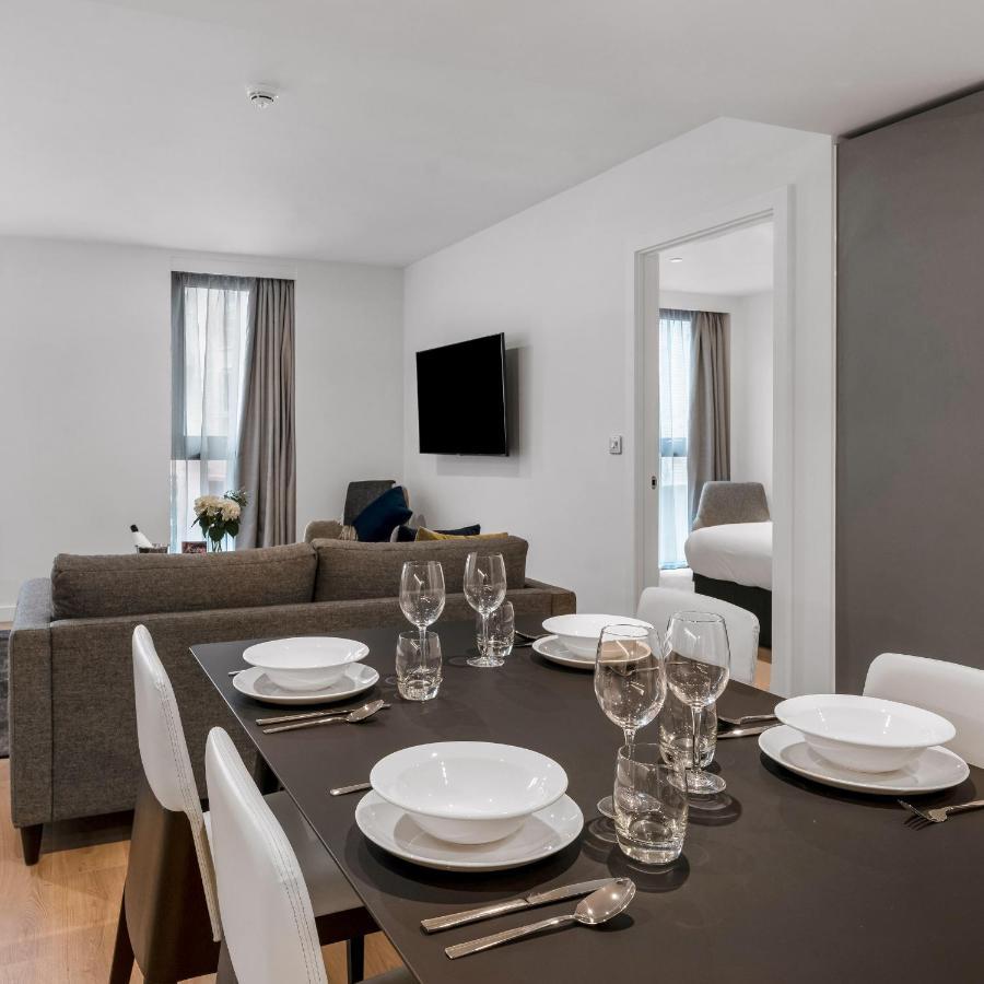 Citysuites 2 Aparthotel Манчестер Екстер'єр фото The photo shows a modern, well-lit living space that features a dining area and a sitting area. In the foreground, there is a dining table set for six, with white plates and crystal glasses arranged neatly. The table is surrounded by white chairs. 

