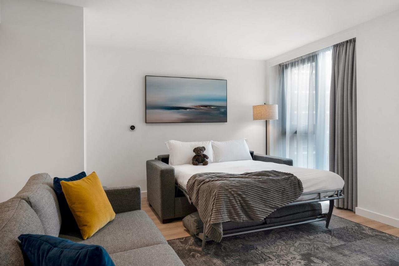 Citysuites 2 Aparthotel Селфорд Екстер'єр фото The photo shows a modern and minimalistic guest room. There is a sofa on the left side, adorned with a colorful cushion. In the middle is a bed that appears to be a pull-out style, featuring white bedding and a gray throw blanket. A stuffed toy is pl