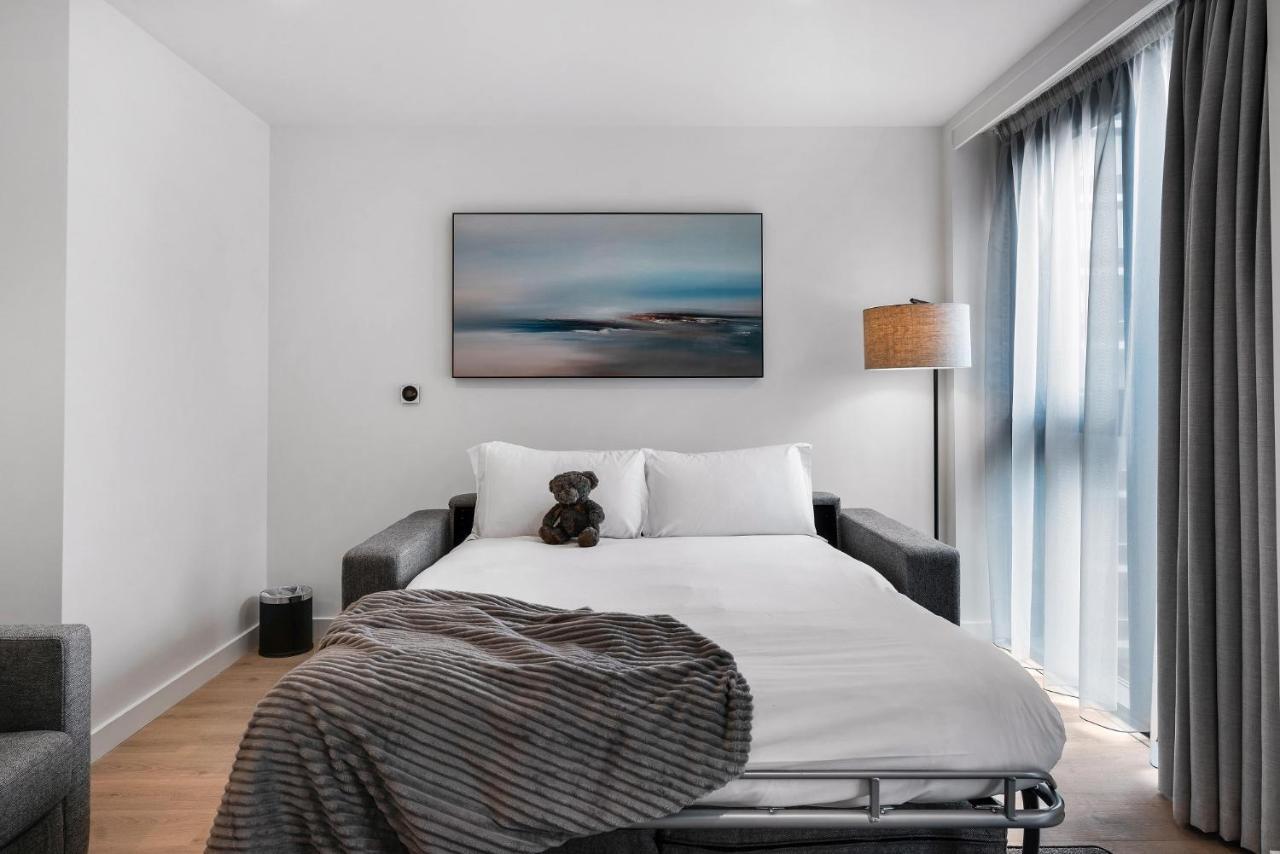 Citysuites 2 Aparthotel Манчестер Екстер'єр фото The photo shows a modern bedroom featuring a bed with white bedding and a decorative throw blanket. There is a teddy bear placed on the bed. A piece of abstract art is hanging on the wall above the bed. The room has a large window with sheer curtains