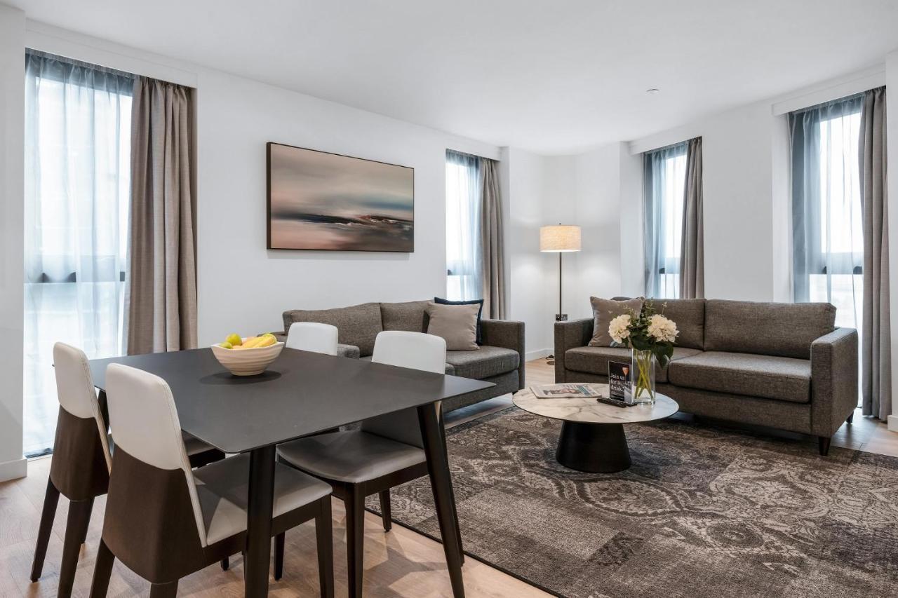Citysuites 2 Aparthotel Селфорд Екстер'єр фото The photo depicts a modern and stylish interior of a living space. On the left, there is a dining table with four chairs, featuring a bowl of fruits on the table. In the background, there's a cozy seating area with a gray sofa and a smaller round cof