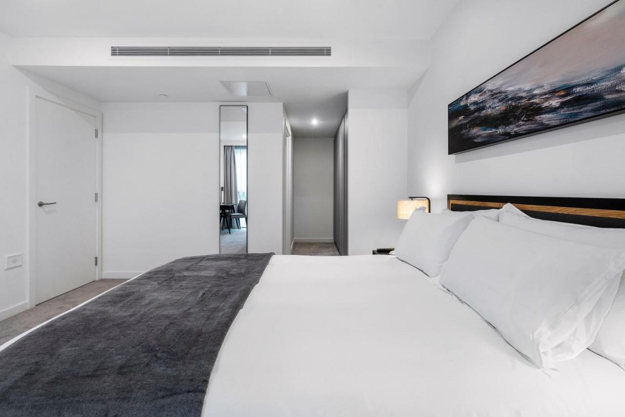 Citysuites 2 Aparthotel Манчестер Екстер'єр фото The photo shows a modern bedroom with a minimalistic design. In the foreground, there is a neatly made bed with white linens and a dark gray throw blanket. To the side, there is a bedside lamp and a piece of abstract artwork on the wall above the bed