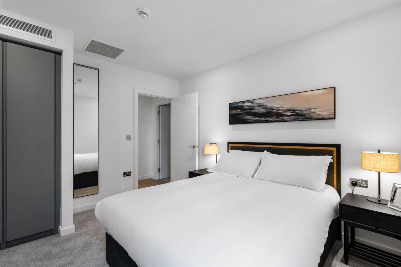 Citysuites 2 Aparthotel Манчестер Екстер'єр фото The photo shows a modern bedroom featuring a large double bed with white bedding. There are two bedside tables with lamps on either side of the bed. The walls are painted white, and there's a piece of artwork above the bed that depicts a landscape. A