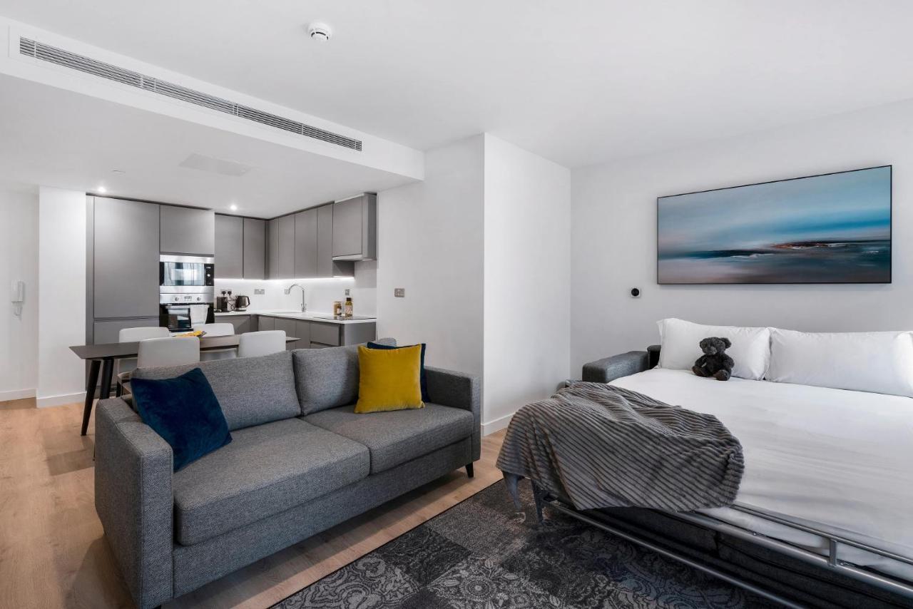 Citysuites 2 Aparthotel Манчестер Екстер'єр фото The photo shows a modern, open-concept living space that includes a kitchen and a sleeping area. 

- **Living Area**: On the left, there's a gray sofa with a blue and yellow throw pillow. A blanket is draped over one side, and there's a small dark ru
