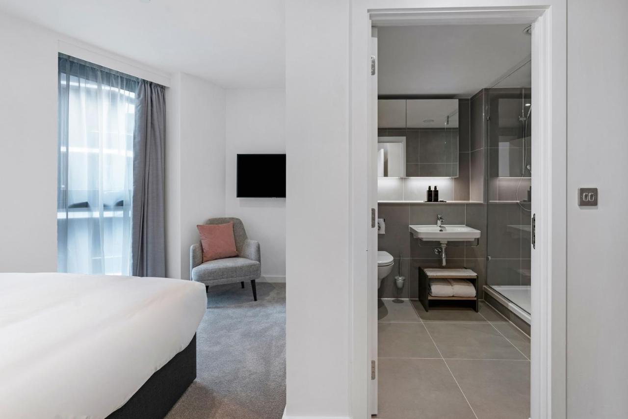 Citysuites 2 Aparthotel Манчестер Екстер'єр фото The photo shows a modern hotel room and bathroom setup. On the left, there is a bed with white bedding and a small sitting area that includes a gray chair and a pillow in a subtle pink shade. A flat-screen TV is mounted on the wall, providing enterta