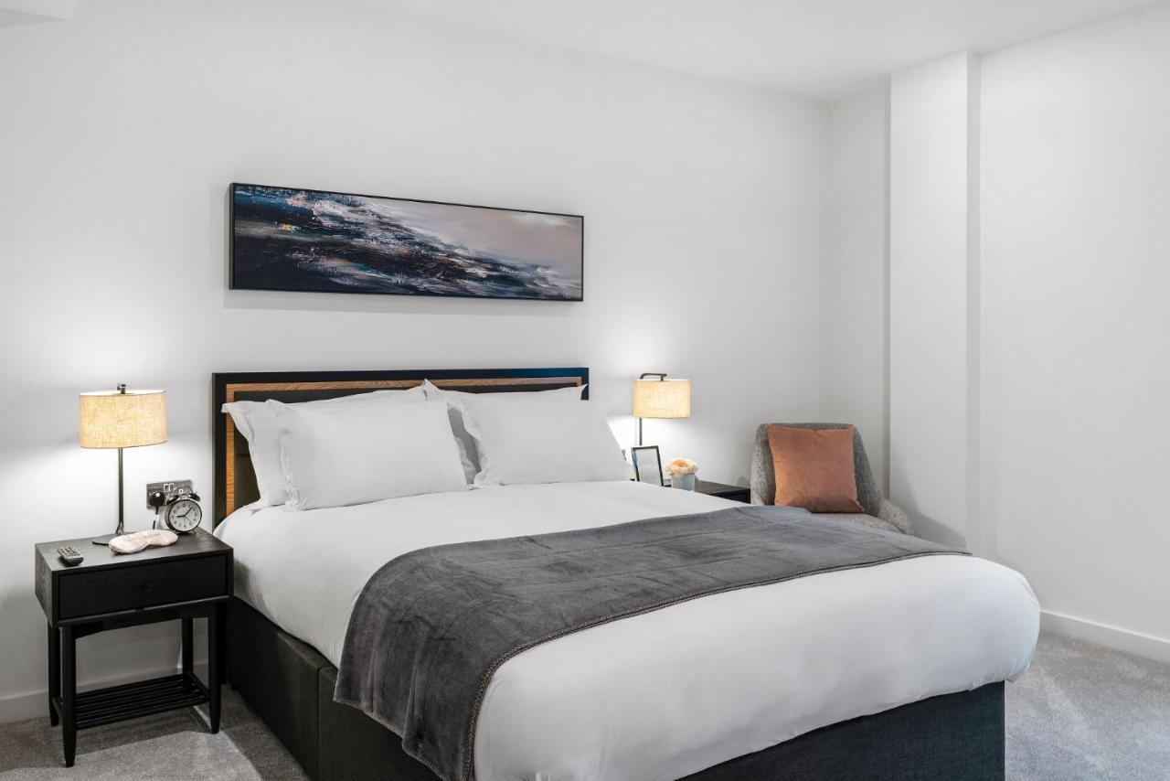 Citysuites 2 Aparthotel Селфорд Екстер'єр фото The photo shows a modern bedroom featuring a large bed with crisp white linens and a grey throw blanket. On either side of the bed, there are bedside tables with lamps. A piece of abstract art hangs above the headboard, and there is a chair in a corn
