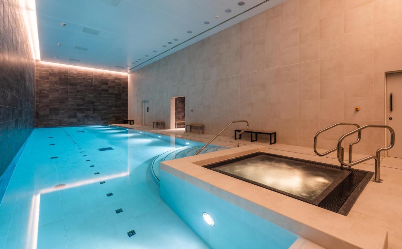 Citysuites 2 Aparthotel Селфорд Екстер'єр фото The photo shows an indoor swimming pool area. The pool is a bright blue color and features smooth, tiled surfaces. On one side, there is a hot tub area with steps leading into it. The walls are made of light-colored stone, and there is ambient lighti