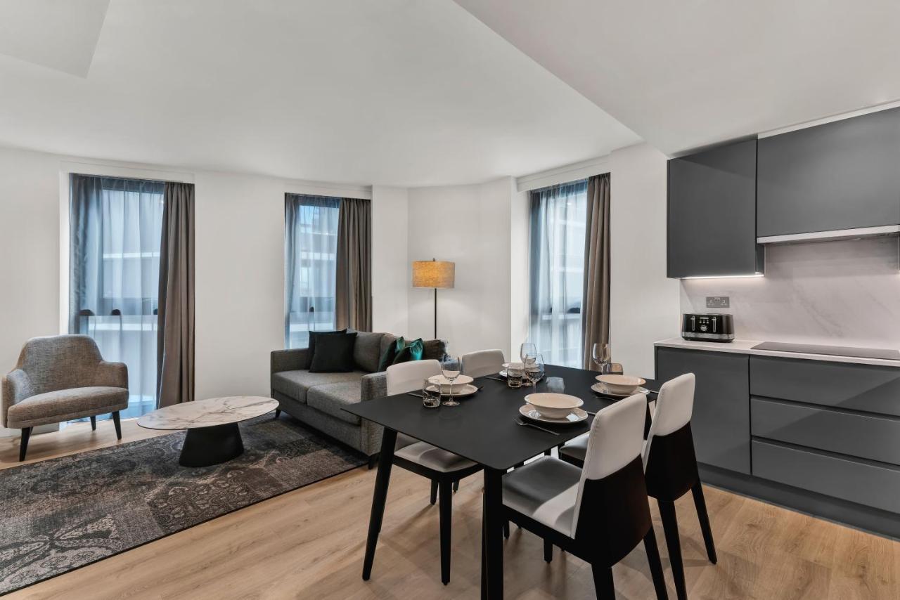 Citysuites 2 Aparthotel Манчестер Екстер'єр фото The photo shows a modern living space that includes a dining area and a seating area. There's a sleek black dining table set with plates and cutlery, accompanied by white and black chairs. To one side, a comfortable gray sofa faces the dining area, c