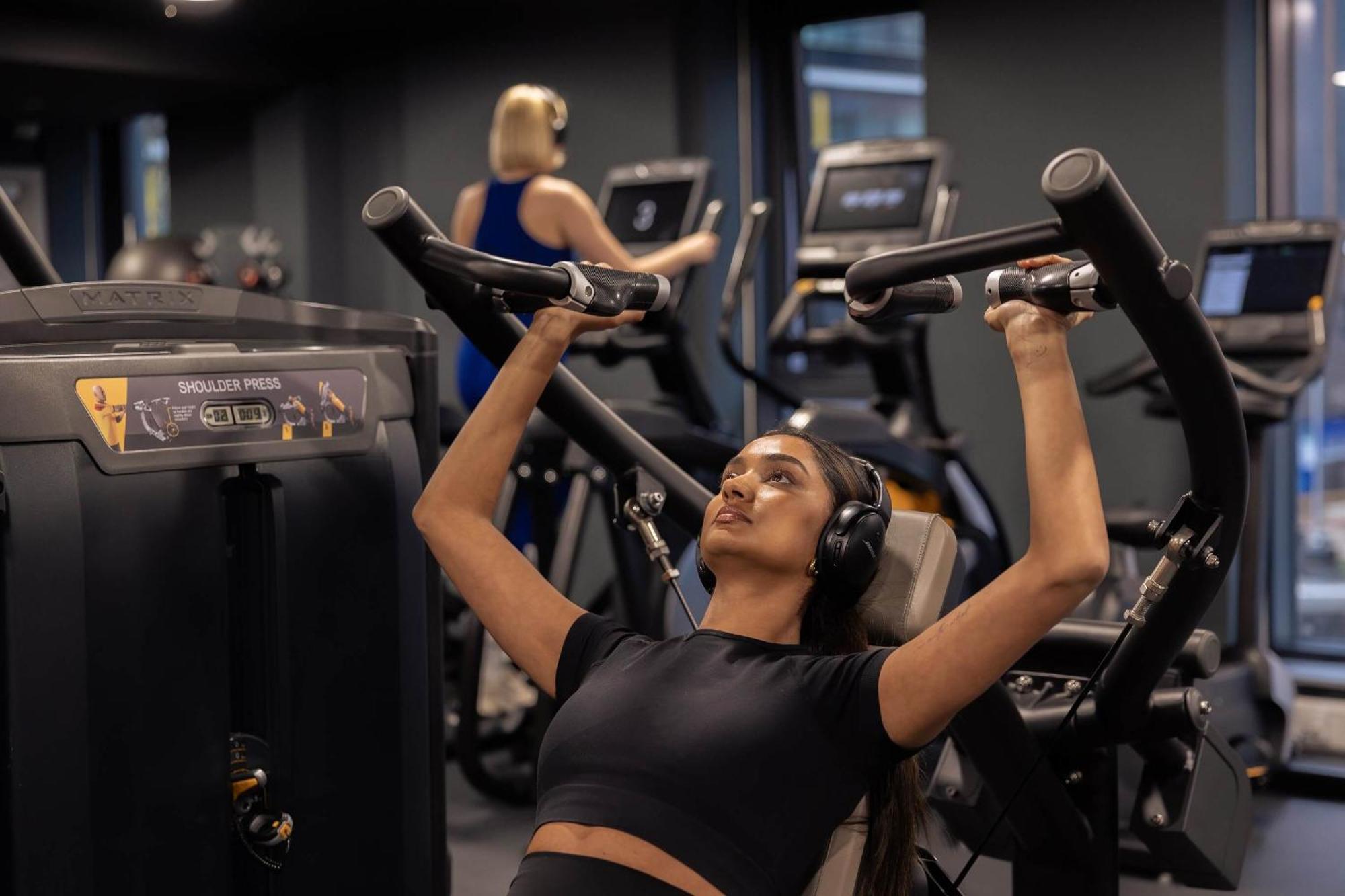 Citysuites 2 Aparthotel Манчестер Екстер'єр фото The photo shows a gym setting with two individuals engaged in physical exercise. In the foreground, a woman is using a chest press machine while wearing headphones, focusing on her workout. She appears to be in a seated position, pushing the handles 