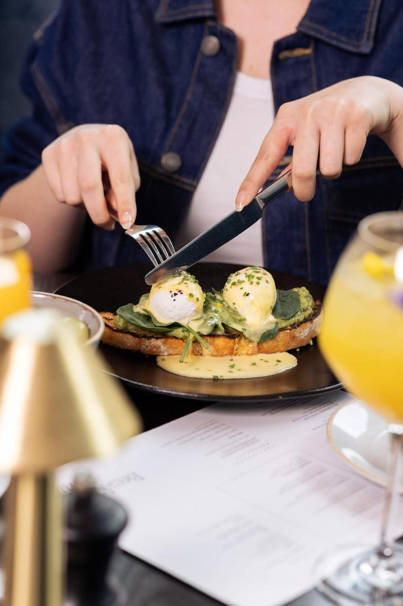 Citysuites 2 Aparthotel Селфорд Екстер'єр фото The photo shows a person preparing to eat a dish of eggs Benedict. They are holding a knife and fork, ready to cut into the meal, which features poached eggs on top of a slice of toast, accompanied by spinach and a creamy hollandaise sauce drizzled o