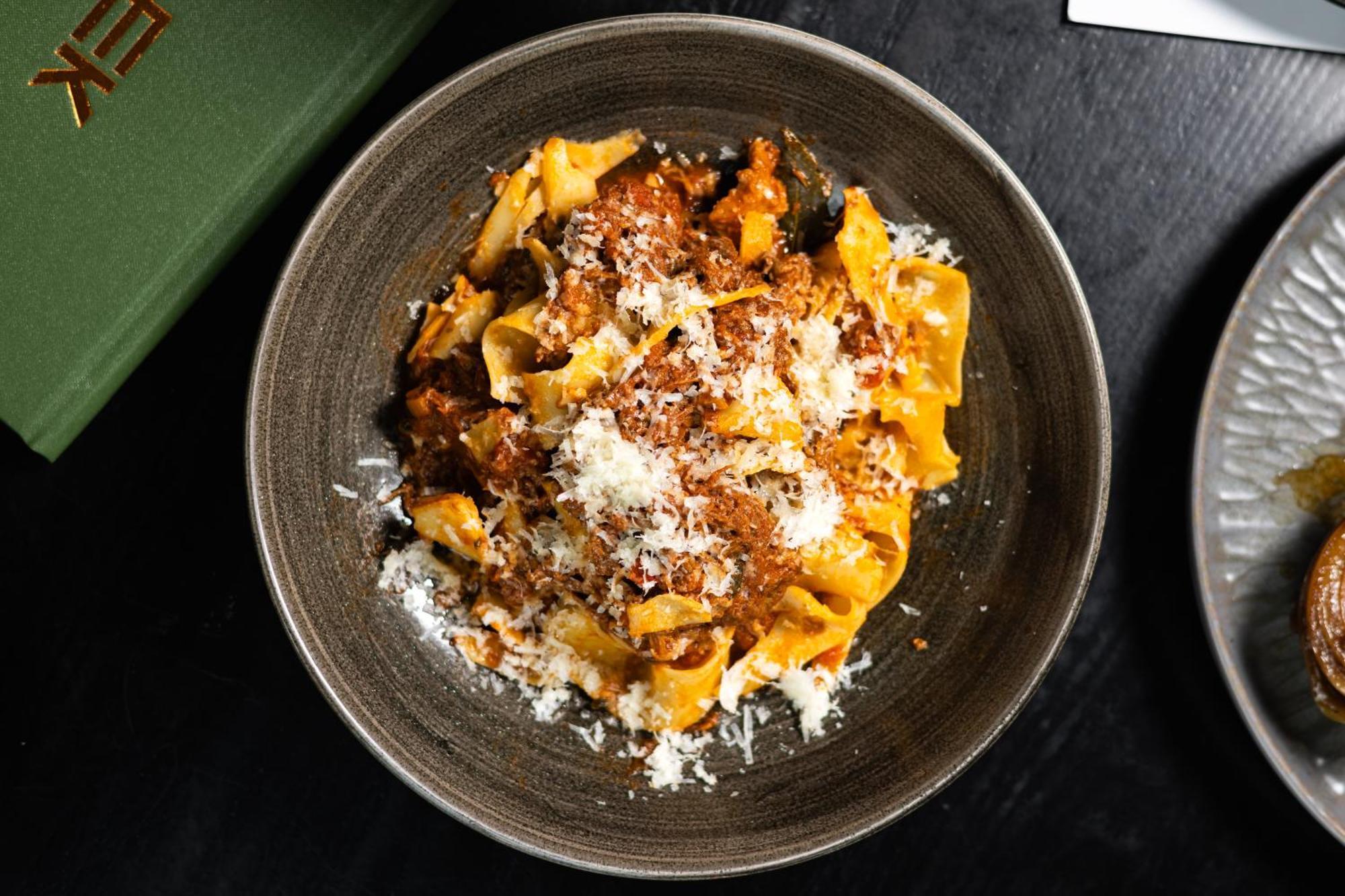 Citysuites 2 Aparthotel Манчестер Екстер'єр фото The photo shows a bowl of pasta featuring wide, ribbon-like noodles, likely pappardelle, fully coated in a rich meat sauce. The dish is garnished with a generous sprinkling of grated cheese, adding a creamy texture to the presentation. The dark bowl 